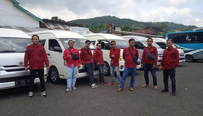 Tim driver rental hiace purwokerto profesional berkerah merah dari BAG Transport Purwokerto berpose ramah di depan unit Toyota Hiace terbaru di garasi pool.