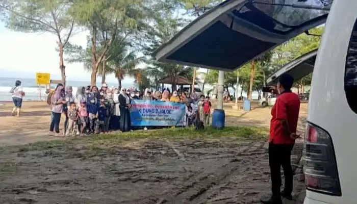Rombongan wisatawan bahagia berfoto bersama di pantai dengan spanduk acara, didampingi driver BAG Transport berkaos merah, menggunakan unit Toyota Hiace terbaru.